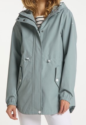 Femme portant une veste de pluie à capuche bleu clair avec fermeture éclair à l'avant, boutons-pression, cordons réglables à la taille et deux poches à rabat sur le devant.