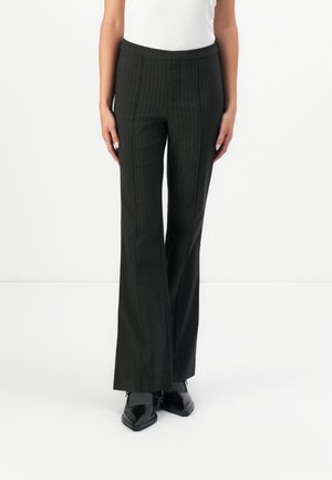 Kvinne iført svarte pinstripe dressbukser, hvit ermeløs topp og svarte spisse sko, stående mot en hvit bakgrunn.