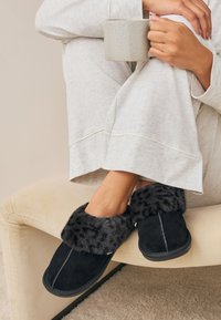 Chaussons en suède noir avec un bord en fausse fourrure douce, présentant un design simple et des coutures contrastantes. Portés avec un pantalon de détente gris clair.