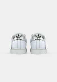 Baskets basses en cuir blanc avec des accents noirs, présentant une surface texturée, un bout arrondi et un logo emblématique à l'arrière.