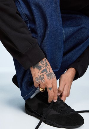 Czarne zamszowe sneakersy z czarnymi sznurowadłami, noszone przez rękę z tatuażami, w tym pierścionkiem na jednym palcu. W tle widoczne ciemnoniebieskie dżinsy.