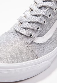 Baskets argentées scintillantes avec une tige texturée, accent en cuir blanc et lacets gris. Elles présentent une semelle extérieure en caoutchouc blanc avec un motif texturé.