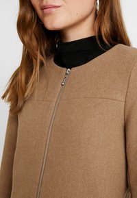 Hnědá džínová bunda na zip s kulatým výstřihem, texturovaná tkanina podobná vlně, se stříbrnými zipovými detaily, kombinovaná s černým rolákem pod ní.