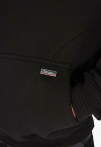 Nahaufnahme einer schwarzen Sweatshirt-Tasche mit geripptem, leicht elastischem Ärmel und einem kleinen rechteckigen silbernen Metall-Logoanhänger, auf dem "Dropstizz" steht.