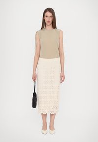 Haut beige sans manches avec détail à nouer, associé à une jupe midi blanche en dentelle avec des motifs circulaires et des bords festonnés, et un sac noir.