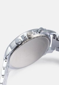 Ρολόι από ανοξείδωτο ατσάλι με στρογγυλή κάσα, γυαλιστερό φινίρισμα και τρία κουμπιά. Διαθέτει βραχιόλι από αλυσίδα και λεία, κενή πίσω όψη.