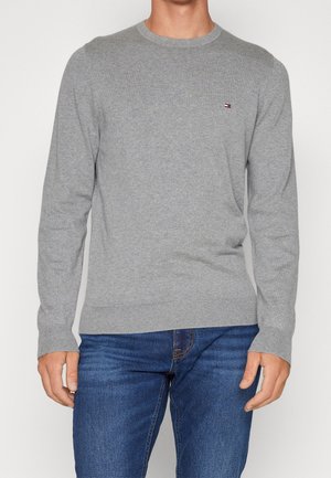 Mężczyzna noszący jasnoszary sweter z okrągłym dekoltem oraz małym logo Tommy Hilfiger na piersi, w połączeniu z niebieskimi dżinsami.