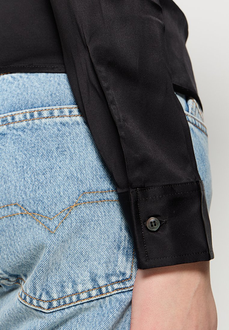 Schwarze Satin-Hemd mit langen Ärmeln und Knopfdetails an den Manschetten, kombiniert mit hellblauen Denim-Jeans mit kontrastierenden Nähten und Design der Gesäßtaschen.