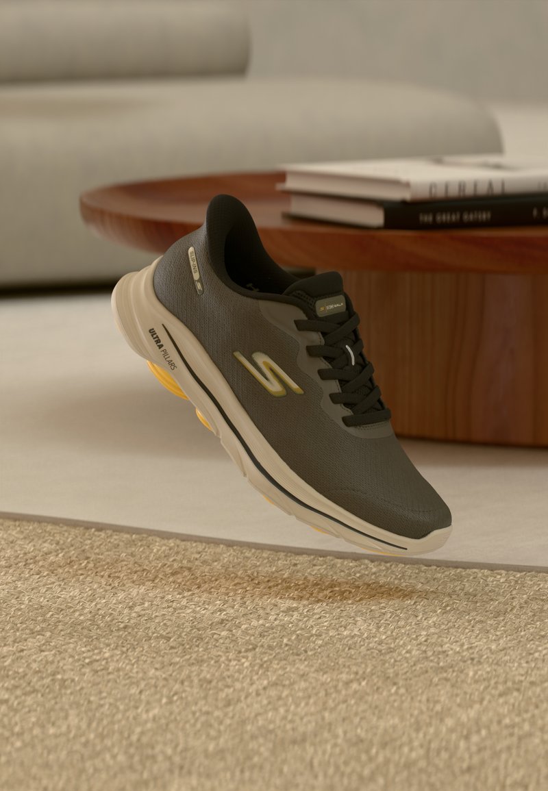 Μαύρο και γκρίζο αθλητικό παπούτσι με μπεζ σόλα και κίτρινες λεπτομέρειες, αιωρούμενο πάνω από μπεζ χαλί κοντά σε ξύλινο τραπέζι με στοιβαγμένα βιβλία.