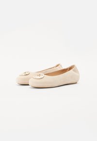 Ballerines en daim beige avec un bout rond, une semelle plate et un accent logo doré à l'avant. Texture lisse sans motifs visibles.