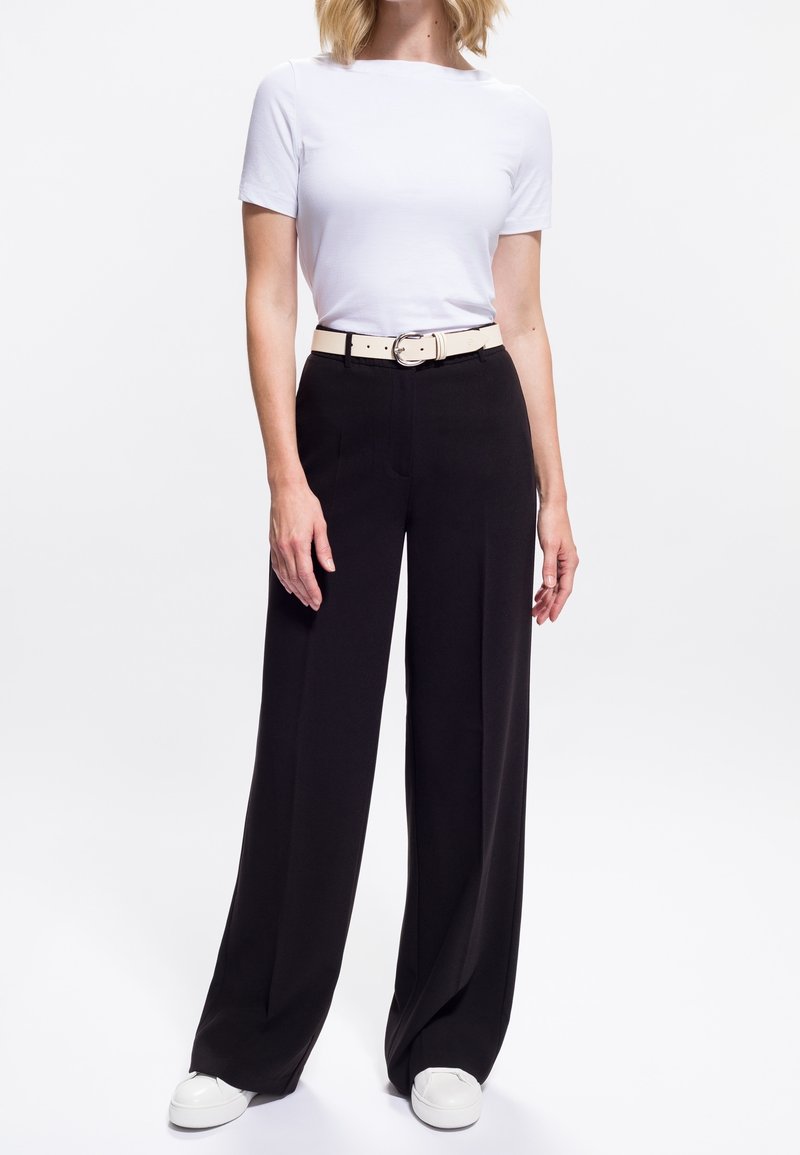 Femme portant une chemise blanche à manches courtes, un pantalon noir taille haute à jambes larges avec une ceinture beige, et des baskets blanches sur fond blanc.