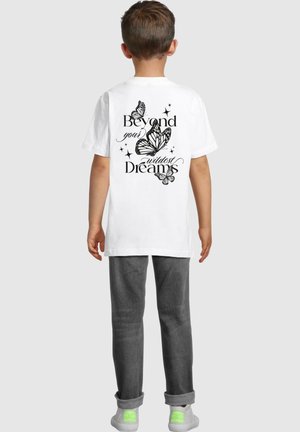 White cotton t-shirt featuring black butterfly graphics and text reading "Beyond your wildest Dreams" on the back. Gray pants and green-accented shoes.