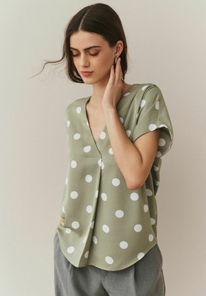Femme aux longs cheveux foncés portant un chemisier vert olive à pois et un pantalon gris, touchant sa boucle d'oreille d'une main.