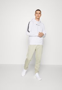 Felpa bianca con cappuccio e logo nero, con strisce sulle maniche, abbinata a pantaloni jogger beige chiaro e sneakers bianche. Design e vestibilità semplici.