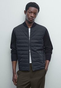 Giubbotto puffo nero con zip frontale e maniche a coste nere, caratterizzato da un design trapuntato e un colletto alto. Indossato sopra una camicia bianca.