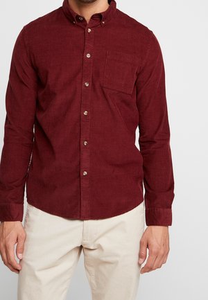 Mężczyzna ubrany w długą, zapinaną na guziki, bordową koszulę z aksamitu sztruksowego z przednią kieszenią oraz beżowe spodnie, stojący na jednolitym tle.