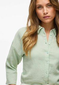 Camicia button-up verde chiaro realizzata in tessuto texturizzato. Presenta un colletto, maniche lunghe e bottoni azzurri chiari.
