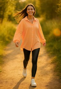 Camicia button-up color pesca con maniche lunghe, abbinata a leggings neri e sneakers bianche, che cammina su un sentiero di terra circondato da vegetazione.