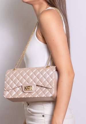 Vrouw draagt een witte top en broek en heeft een beige doorgestikte handtas met gouden kettingriem op haar schouder.