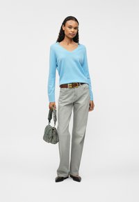  világoskék V nyakú pulóver, szürke egyenes szárú farmernadrág, barna öv és egy textúrált zöld kézitáska; a modellen fekete hegyes orrú lapos cipők vannak.