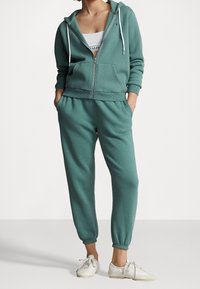 Grön zip-up hoodie i mjukt material med framfickor och dragsko. Sammanlagt med matchande tapered joggers och vita sneakers.