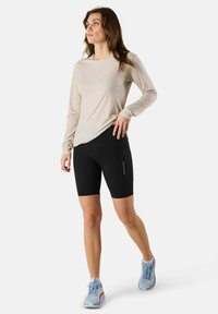 Lichtbeige lange mouwtop, zwarte getailleerde shorts met een ritszak, en blauwe sportschoenen met oranje accenten. Gladde stoftextuur.