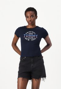 Navy mėlyna įgulos marškinėliai su trumpomis rankovėmis, kuriuose yra balta ir raudona grafika su tekstu "Original Tommy Jeans New York", įkomponuotu centre. Derinami su juodais džinsiniais šortais.
