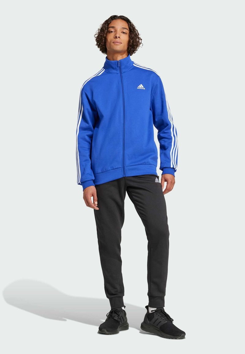 Blå sportjacka med dragkedja och vita randiga detaljer, ihop med svarta träningbyxor och svarta sneakers. Materialet ser mjukt och flexibelt ut.