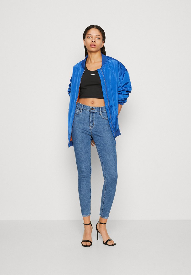 Blaue Bomberjacke, schwarzes Croptop und enge blaue Jeans. Merkmale sind das hochgeschnittene Design, die glatte Textur und schwarze Riemchen-Heels.