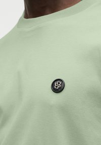 Ανοιχτό πράσινο βαμβακερό μπλουζάκι με ριμπ λαιμόκοψη και στρογγυλή μαύρη ετικέτα στο στήθος που φέρει ασημί έμβλημα.