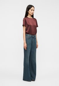 Μπορντό σατέν τοπ με κοντά μανίκια, συνδυασμένο με ψηλόμεσα, φαρδιά μπλε τζιν παντελόνια. Μινιμαλιστικός σχεδιασμός με λεία υφή.
