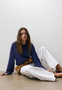 Donna con lunghi capelli castani seduta sul pavimento, indossa una maglietta a maniche lunghe a righe blu e nere, pantaloni bianchi e scarpe rosse, davanti a una parete bianca semplice.