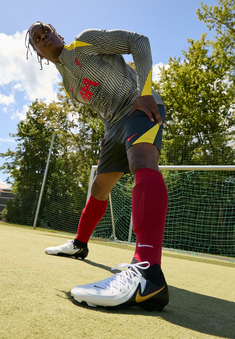 Maglia da football Nike con strisce bianche e nere, dettagli gialli, abbinata a pantaloni scuri e calzettoni rossi. Presenta scarpe chiare e nere con uno swoosh dorato.