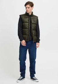 Wattierte Weste in dunklem Olivgrün, mit hohem Kragen, Reißverschluss an der Vorderseite und gestepptem Muster. Getragen über einem schwarzen Langarmshirt und blauen Jeans.