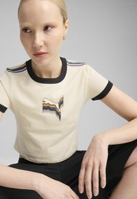 Kremfarvet cropped t-shirt med sorte accenter, prydet med en regnbue-stribet design på skuldrene og en broderet grafik på forsiden.