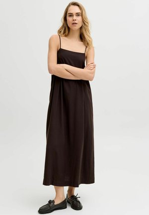 Lyshåret kvinde stående med armene over kors iført en ærmeløs sort maxi-kjole og sorte loafers på en ensfarvet hvid baggrund.