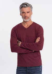 Camiseta de manga larga y escote en V de color burdeos. Textura suave, diseño ajustado, con los brazos cruzados, combinada con jeans azules desgastados.