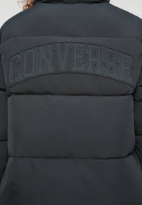 Μαύρο μπουφάν τύπου puffer, με ανάγλυφο λογότυπο "CONVERSE" από καουτσούκ στην πλάτη. Σχεδιασμένο με ραμμένα τμήματα και λείο φινίρισμα.