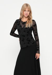 Schwarzes langärmeliges Oberteil mit floraler Struktur, durchsichtigen Ärmeln, V-Ausschnitt, kombiniert mit einem fließenden schwarzen Rock. Weicher, leichter Stoff.