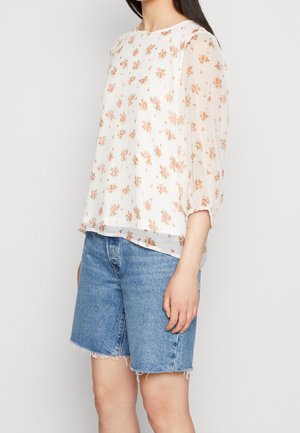 Camicia bianca leggera e trasparente con motivo floreale nei toni del rosa e del verde. Vestibilità rilassata, maniche a palloncino, abbinata a pantaloni di jeans corti.