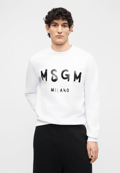 Νεαρός με σγουρά μαλλιά φοράει λευκό φούτερ με το κείμενο "MSGM MILANO" και μαύρο παντελόνι, στέκεται μπροστά σε μονόχρωμο λευκό φόντο.