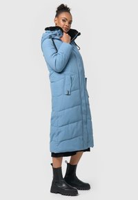 Blue long padded coat with a hood, featuring a black faux fur collar, front pockets, and a quilted pattern. Worn with black ankle boots.