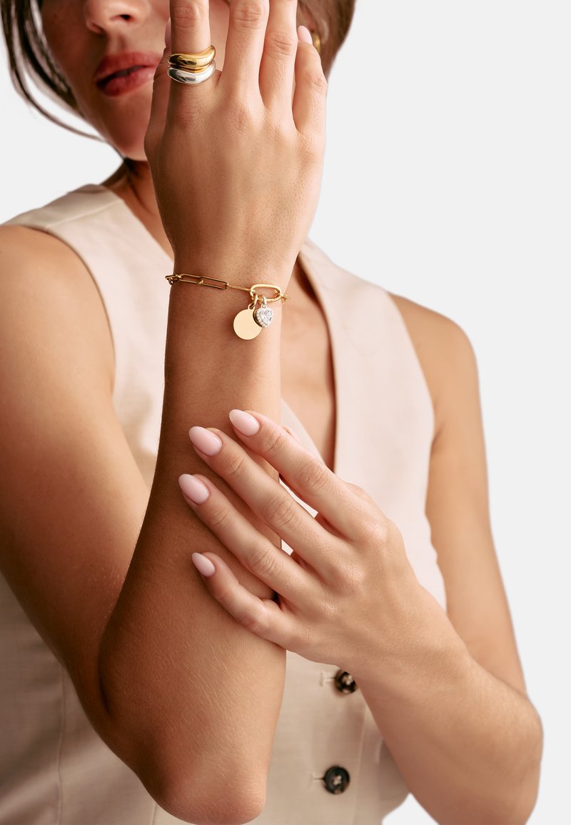 Goldene Kettenarmband mit einem Herzanhänger und zwei runden Anhängern; glatte Textur; präsentiert an einem Handgelenk mit manikürten Händen.