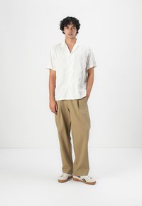 Short-sleeve white shirt with embossed leaf pattern, paired with loose beige trousers and white sneakers featuring black stripes.