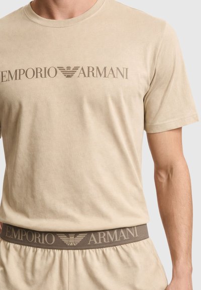 Μπεζ κοντομάνικη μπλούζα Emporio Armani και αντίστοιχα κάτω μέρη με λογότυπο και έμβλημα αετού στο στήθος και τη ζώνη μέσης.