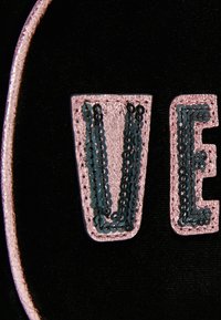 Tela negra con letras cosidas brillantes en rosa "V" y "E", detalladas con lentejuelas negras contra un borde circular rosa.