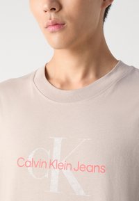 T-shirt à col rond beige clair fabriqué en coton doux, arborant un grand logo "CK" blanc et le texte "Calvin Klein Jeans" en rose vif.