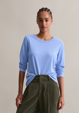Junge Frau mit geflochtenem Haar, die ein hellblaues langärmeliges Shirt trägt, das in olivgrüne hochtaillierte Hosen gesteckt ist, vor einem schlichten Hintergrund.