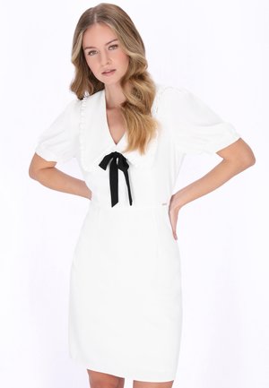 Donna con lunghi capelli biondi ondulati che indossa un abito bianco con maniche a sbuffo e un grande colletto legato con un nastro nero, in piedi con le mani sui fianchi.