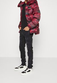 Punainen ja harmaa kuvioitu toppatakki, musta t-paita, kuluneet mustat farkut, joissa on teksturoituja kohtia, ja valkoiset lenkkarit mustilla yksityiskohdilla.